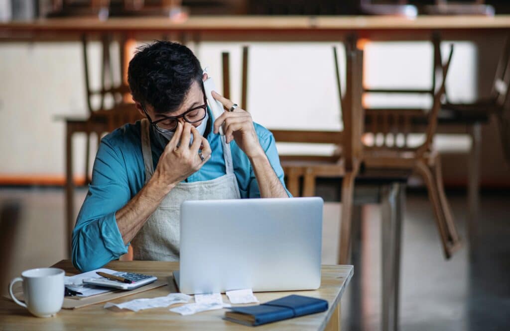 Stressed small business owner in an apron sitting at a desk with receipts, a calculator, and a laptop, struggling to review financial and inventory data