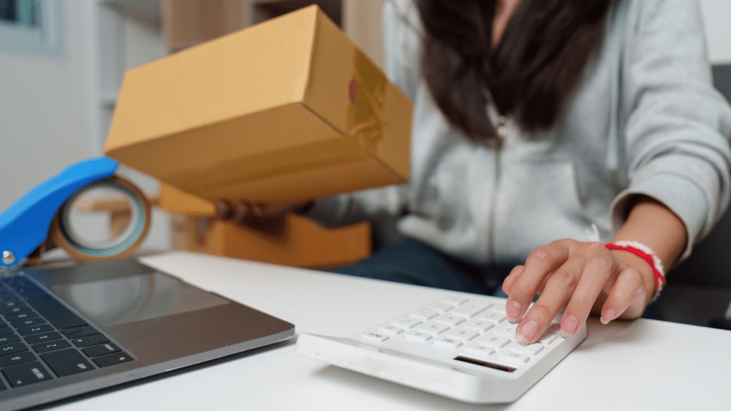Business owner calculating inventory costs with boxes, laptop, and calculator to identify stock management issues.